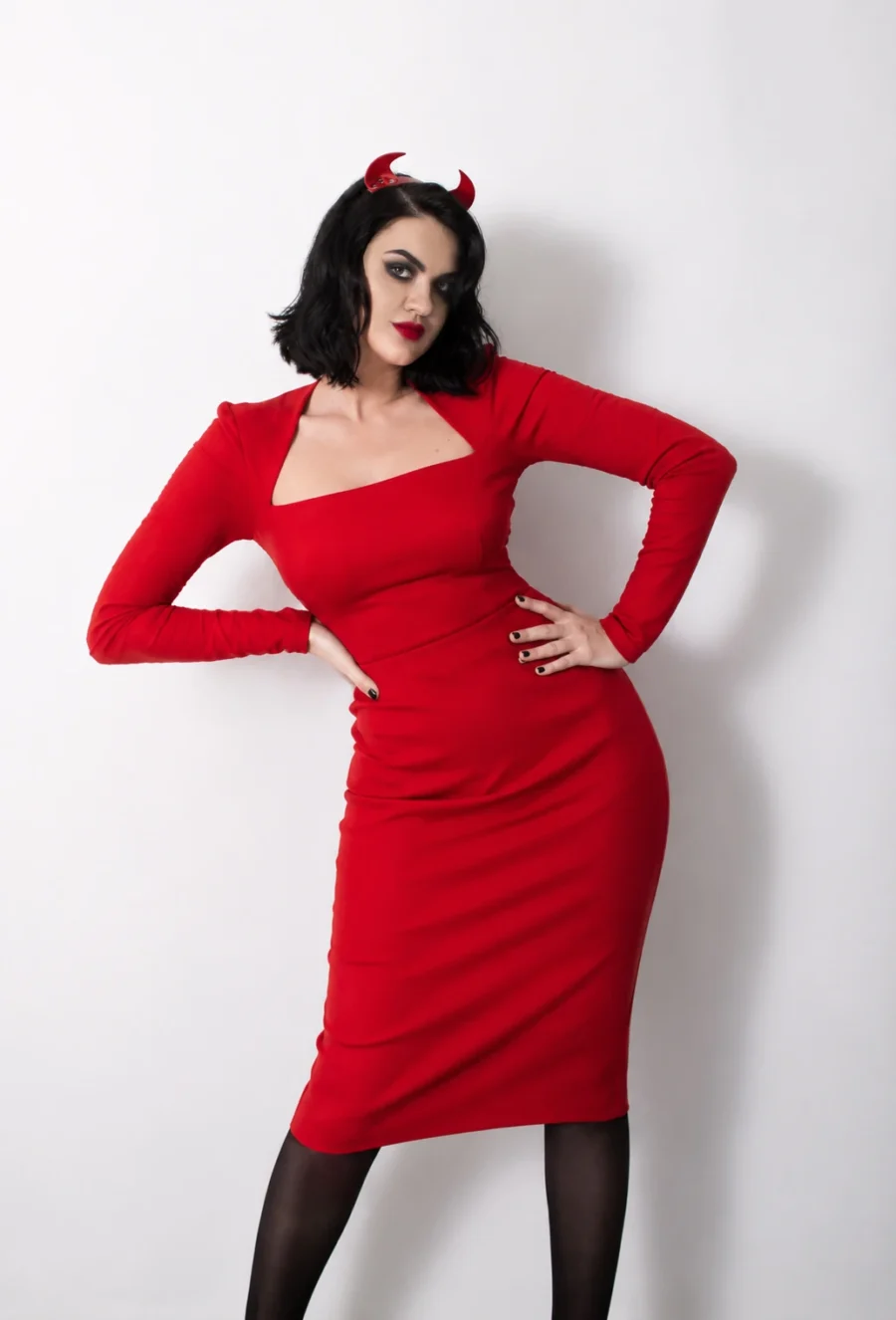Artist Lady Redhorns in a fitted red dress and small red horns, hands on hips against a plain pale backdrop.